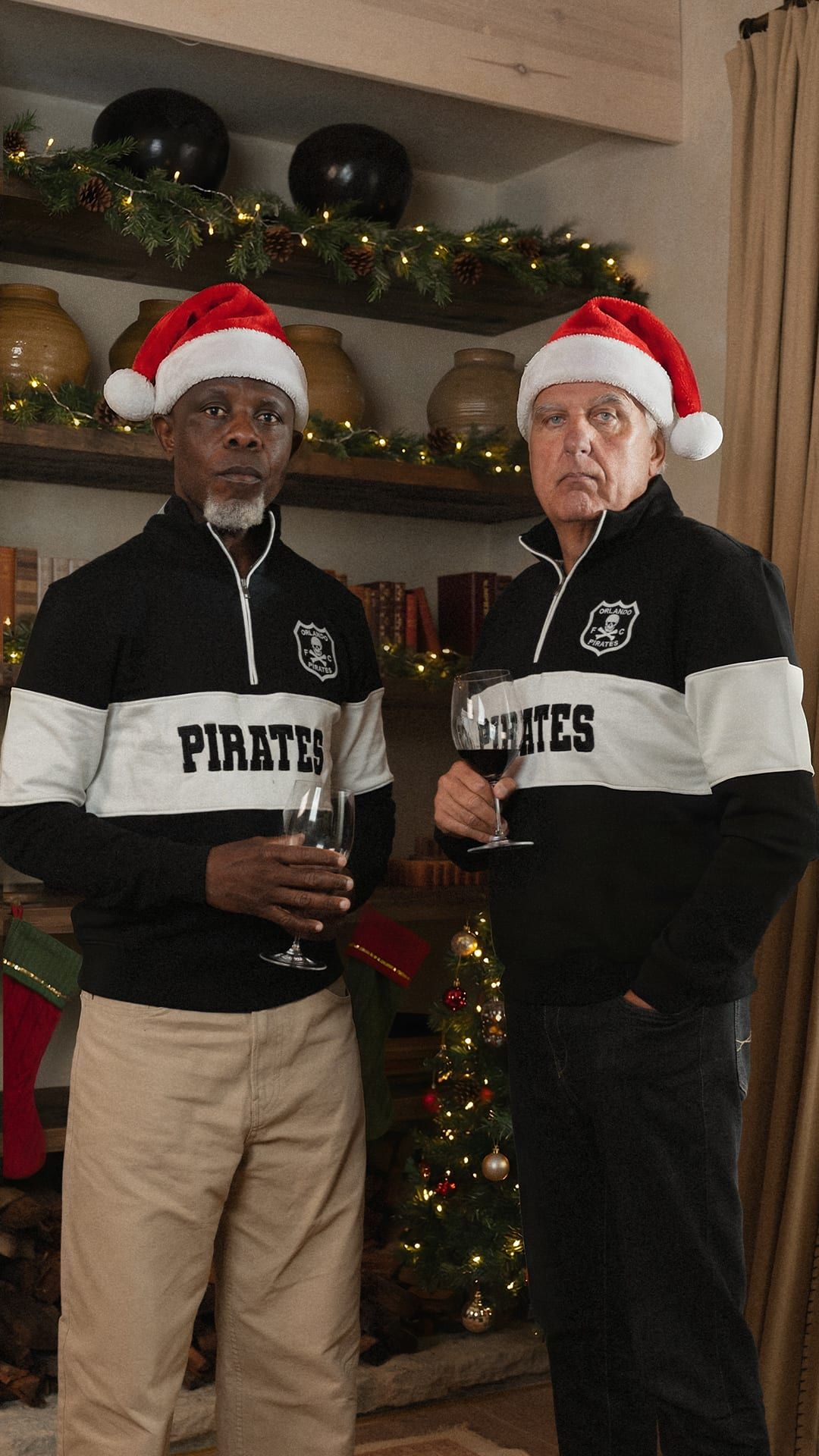 Two men in Pirates quarter-zips and Santa hats by a Christmas tree