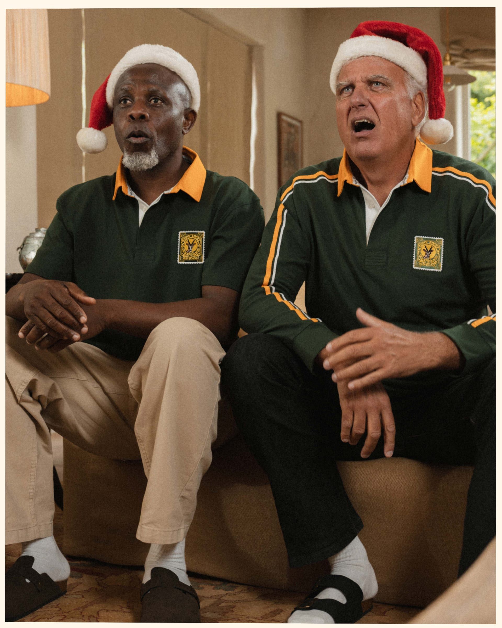 Two men in Springbok polos and Santa hats mid-carol