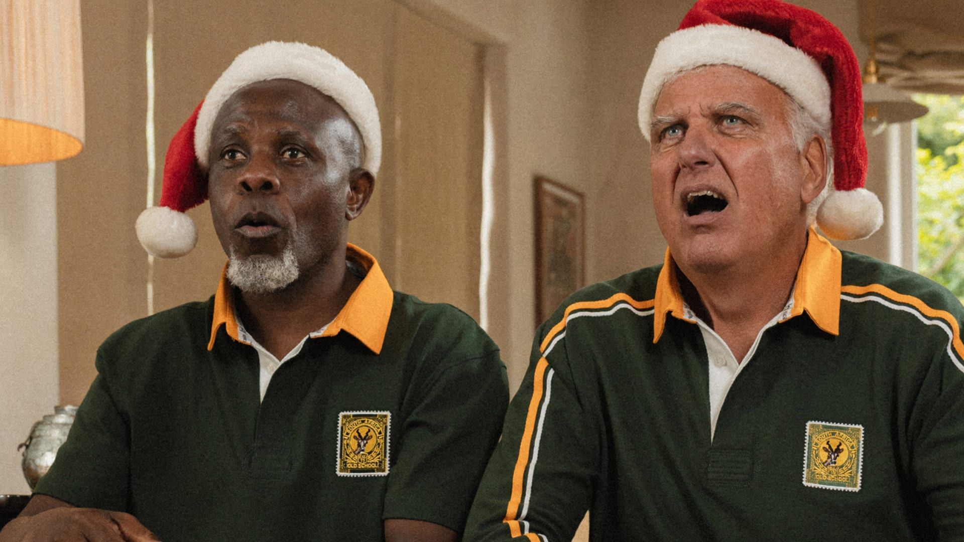 Two men in Springbok polos and Santa hats mid-carol, Old School Christmas gifting
